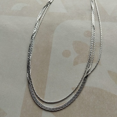 Colar fino prateado com duas correntes entrelaçadas sobre fundo bege com padrão abstrato
