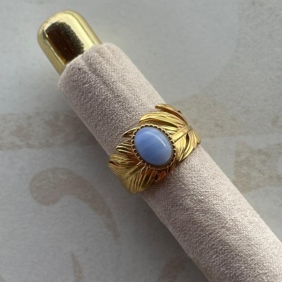Anel dourado com pedra azul claro e suporte almofadado bege.