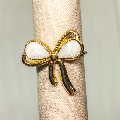 Anel dourado em forma de laço com preenchimento branco, em suporte de tecido bege