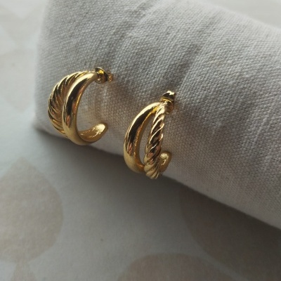Par de brincos dourados em metal com dois designs diferentes em tecido bege.