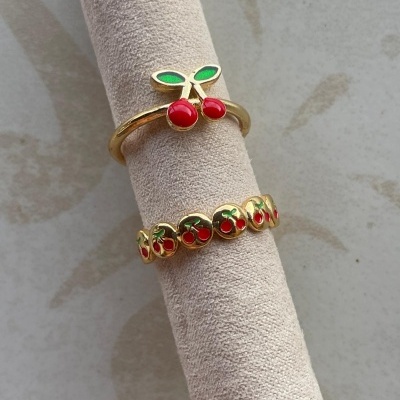 Dois anéis dourados com cerejas vermelhas e folhas verdes num suporte de tecido cor creme.