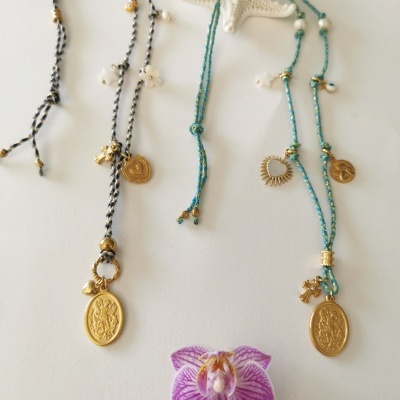 Dois colares com medalhões dourados em cordões preto e branco e verde, com pendentes e pérolas, sobre fundo branco com estrela do mar e flor roxa.