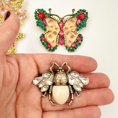 Broche inseto dourado com pedras e abdómen branco numa mão, broche borboleta colorido ao fundo.