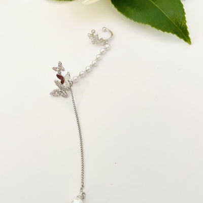 Brinco prateado com borboletas e pérola com flores brancas ao fundo.