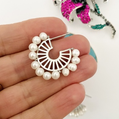 Brinco meia-lua em metal com pérolas, mão segurando, broche de flor ao fundo