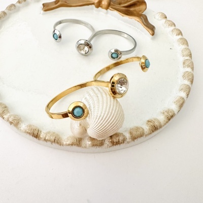 Duas pulseiras, uma dourada e outra prateada, com pedras azuis e transparentes sobre prato branco decorativo.