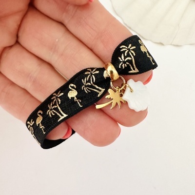Pulseira preta com padrões dourados e pendentes, segurada por mão com unhas rosa