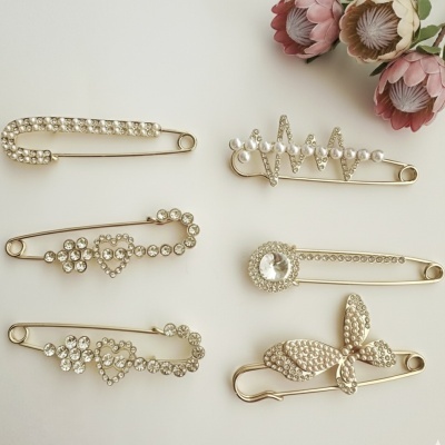 Seis alfinetes de segurança decorativos com pedras e pérolas, designs variados, fundo branco e flores artificiais.