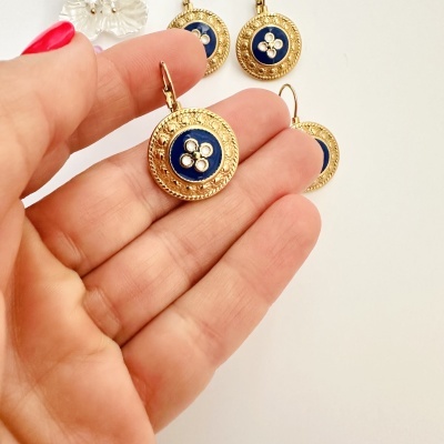 Brincos dourados redondos com detalhe azul no centro sendo segurados por uma mão feminina.