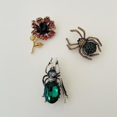 Três broches em forma de flor e aranhas com pedras coloridas e metal sobre fundo branco.