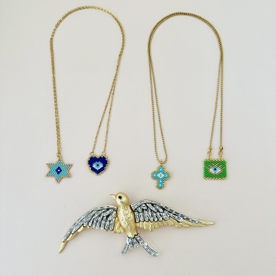 Joias coloridas com correntes douradas e broche de pássaro em fundo neutro.
