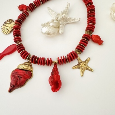 Colar vermelho com pendentes de conchas e estrela dourada sobre fundo branco com conchas naturais.