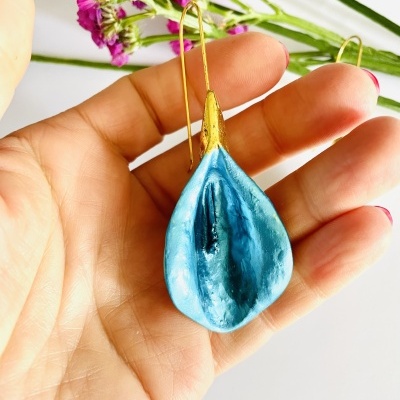 Brinco pendente azul metálico com detalhe dourado segurado em mão com flores ao fundo