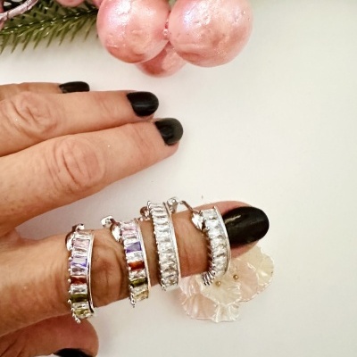 Anéis prateados com pedras coloridas e unhas pretas num fundo branco com decoração de frutos rosa.