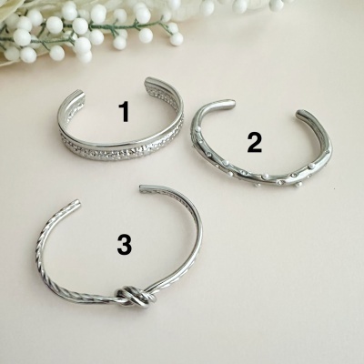 Três pulseiras prateadas com designs texturizados e um nó central na terceira pulseira.