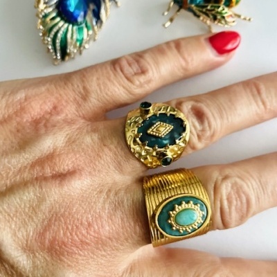 Mão com anéis dourados com pedras azuis e broches decorativos ao fundo.
