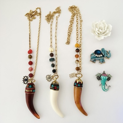 Colares com dentes decorativos, contas coloridas, pendentes de olho, broches de elefante azuis e flor branca sobre fundo branco.