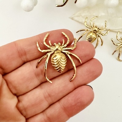 Broche dourado em forma de aranha segurado na mão, com outras aranhas ao fundo