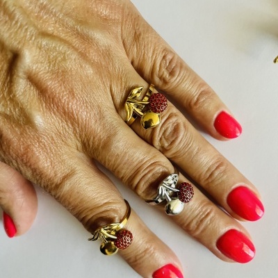 Mão com três anéis de cereja, dois dourados e um prateado, unhas vermelhas.