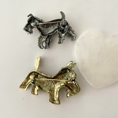 Broches em forma de burros, um prateado e outro dourado, com alfinetes na parte de trás, sobre fundo branco ao lado de coração translúcido