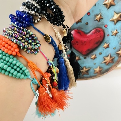 Pulso com várias pulseiras coloridas de contas e franjas diante de placa decorativa com coração e estrelas