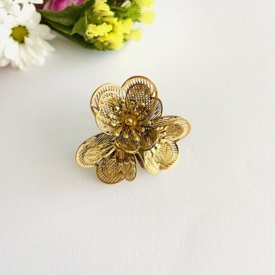 Anel dourado em forma de flor com pétalas detalhadas sobre fundo branco e flores coloridas