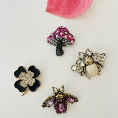Broches decorativos em formas de trevo, cogumelo e abelha com várias pedras e uma flor rosa ao fundo
