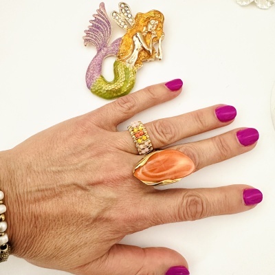 Figura decorativa de sereia com cauda lilás e escamas verdes ao lado de uma mão com unhas pintadas e anéis coloridos