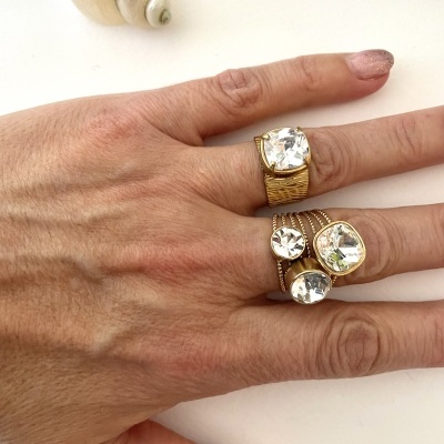 Mão feminina com três anéis dourados com pedras claras e concha ao fundo