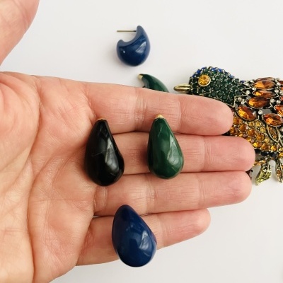 Brincos pendentes em gota preta, verde e azul escuro numa mão, com broche colorido ao fundo num fundo branco.