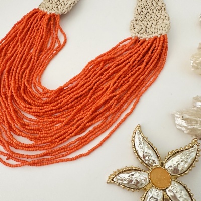 Colar de contas laranja em tecido bege com adereço floral prateado e dourado.