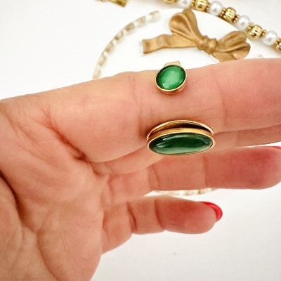 Mão segurando dois anéis com pedras verdes, colar de pérolas e laço dourado ao fundo