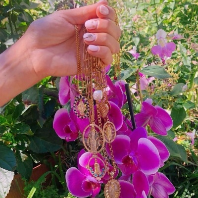 Mão segura colares dourados com pendentes contra flores orquídeas roxas.