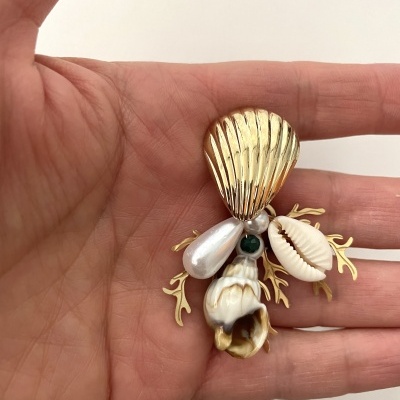 Broche artesanal com conchas naturais, pérola e detalhes dourados mostrado numa mão.