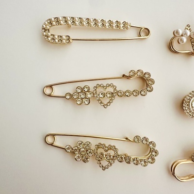 Agulhas de segurança douradas decoradas com cristais e pérolas falsas numa superfície branca.