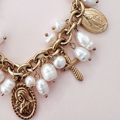 Pulseira dourada com pérolas brancas e pendentes religiosos sobre fundo rosa