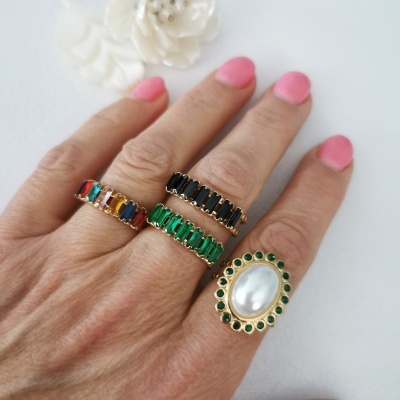 Mão com quatro anéis dourados com pedras coloridas, verdes, pretas e branca, unhas cor-de-rosa, fundo branco com flores decorativas.