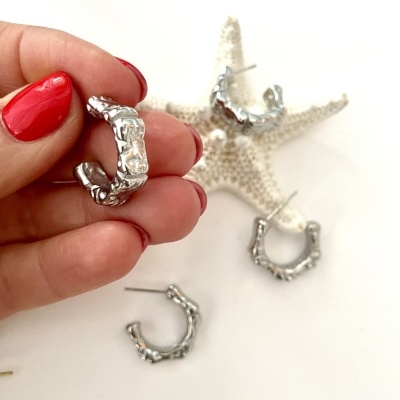 Brincos prateados com textura de ramo exibidos numa mão com unhas vermelhas e estrela-do-mar no fundo