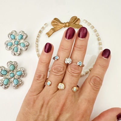 Mãos femininas com anéis de metal e pedras verde-água e cristais em fundo branco com decoração.