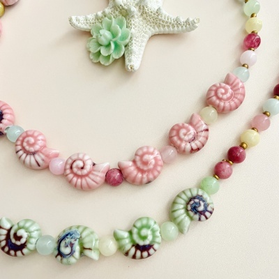 Colares com contas em formato de conchas em rosa e verde com pedras coloridas, sobre fundo branco com estrela-do-mar e suculenta