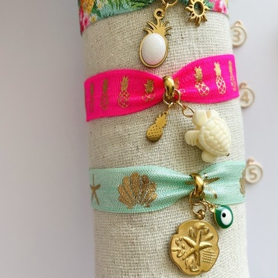 Pulseiras coloridas elásticas com pendentes dourados e brancos, sobre suporte bege cilíndrico.