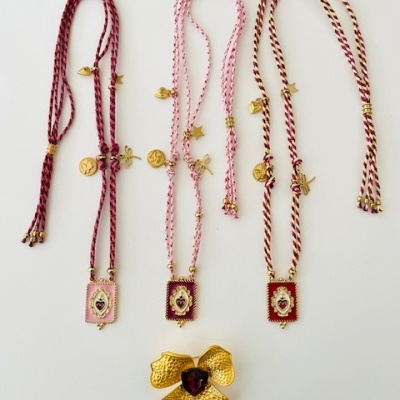 Três colares com cordões trançados e pendentes retangulares decorados, e um broche dourado em forma de flor com pedra vermelha.