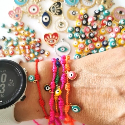 Pulseiras coloridas de fio trançado com olhos turcos, braço com relógio digital, fundo branco com mais amuletos.