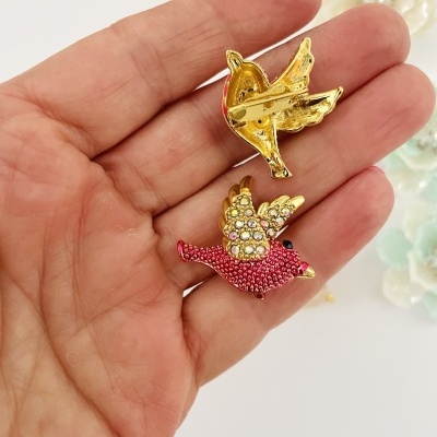 Broches de passarinho em metal dourado com detalhes em strass e esmalte vermelho, segurados numa mão.
