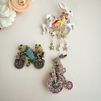 Três broches decorativos com coelhos e sapos em bicicleta e coelho com pendentes de estrelas.
