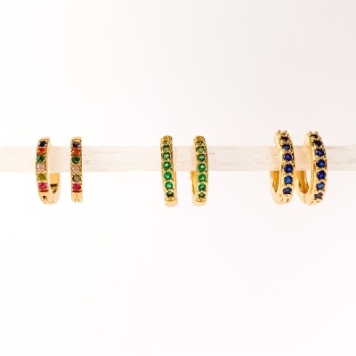 Três pares de brincos argola dourados com pedras coloridas pendurados numa barra.