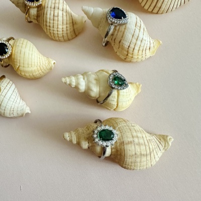 Anéis com pedras azuis e verdes em conchas naturais sobre fundo bege.