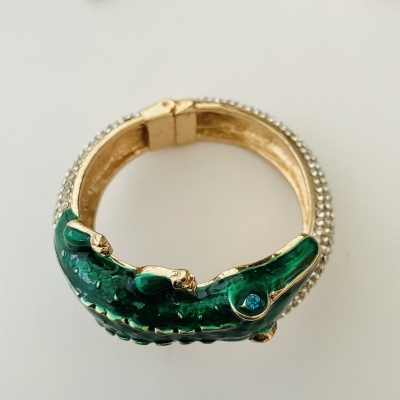 Pulseira dourada com design de crocodilo verde e olho azul sobre superfície branca
