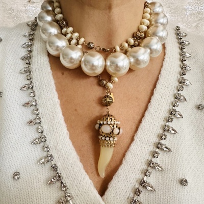 Colar de pérolas grandes e pequenas com pendente dourado em forma de dente, usado sobre camisola branca com bordados prateados