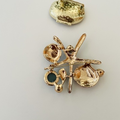 Peça de joalharia dourada com forma abstrata e pedra verde escura sobre fundo branco